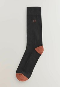 Musta sukka, jossa on poimutettu pintakuvio, kontrastiväriset ruosteenoranssit vahvistetut varvas- ja kantapääosat sekä pieni logo resorin lähellä.