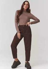Brązowa bluzka z długim rękawem i wysokim kołnierzem, zestawiona z ciemnymi brązowymi spodniami z wysokim stanem. Modelka nosi masywne czarne buty na platformie.