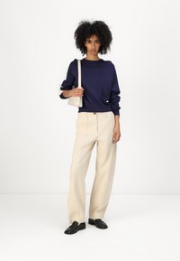 Felpa navy con dettagli testurizzati, pantaloni a gamba larga color crema e scarpe nere. Il modello tiene una piccola borsa tote beige.