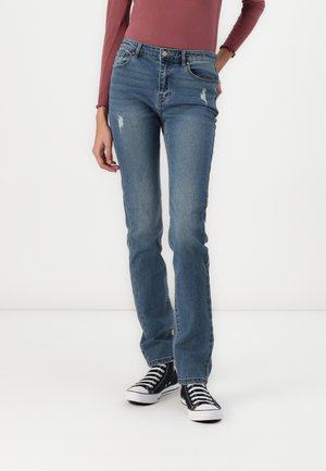 Vrouw draagt vervaagde blauwe rechte jeans met lichte slijtage en zwarte hoge sneakers, staand tegen een witte achtergrond.