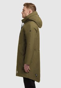 Oliveza zaļa parka ar kapuci, ar rāvējslēdzēju priekšā, sānu kabatām un melnu logo akcentu. Gluda tekstūra un ceļgala garuma dizains.