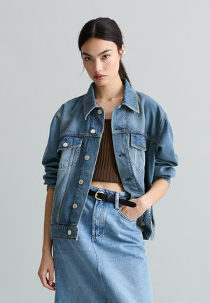 Jonge vrouw met een blauwe spijkerjack en rok, een kort bruin topje en een hand in haar zak staand tegen een effen grijze achtergrond.