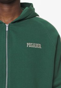 Felpa verde con zip, realizzata in materiale morbido, dotata di un ampio cappuccio, con dettagli grigio chiaro e la scritta "PEGADOR" ricamata in bianco sopra la zip.