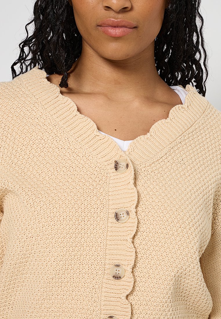 Beige gebreide cardigan met een golvende kraag, vijf knopen en een gemêleerd stof. Eenvoudig ontwerp zonder extra patronen of accenten.