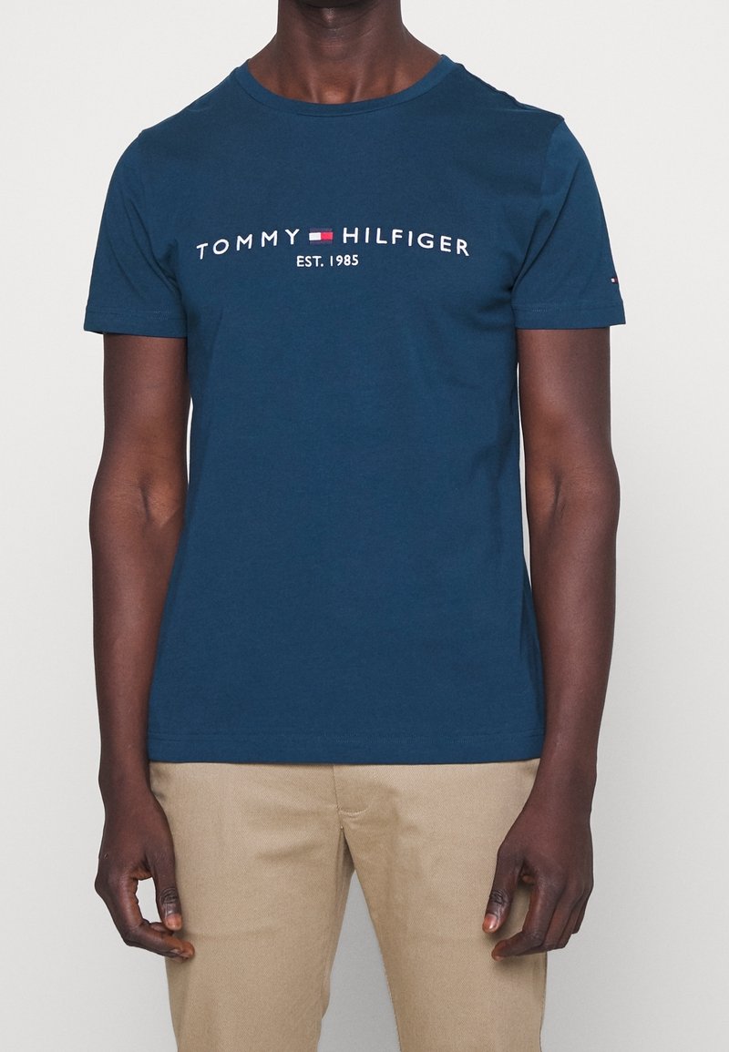 Man med en marinblå Tommy Hilfiger t-shirt med logotyp och texten "EST. 1985", i kombination med beige byxor, som står mot en enfärgad bakgrund.