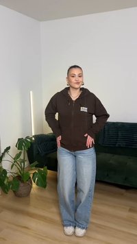 Brun hættetrøje med lynlås og logo, lyseblå vide jeans og hvide sneakers. Baggrunden har en grøn sofa og en potteplante.