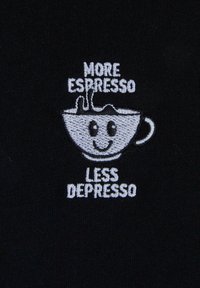 Sort stof med hvid broderi, der viser en smilende kaffe-kop og teksten "Mere Espresso Mindre Depresso" ovenfor og nedenfor koppen.