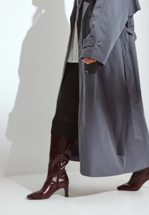 Oseba hodi v bordo usnjenih škornjih, črnih širokih hlačah in dolgem sivi plašču proti enostavni svetli ozadju.