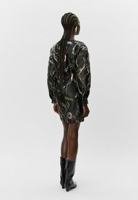 Moteris su ilgomis kasomis, vilkinti tamsų gėlėtą suknelę ir juodus vidutinio ilgumo aukštakulnius batus, stovi prieš vienodą šviesų foną.