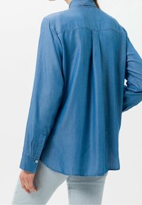 Jasnoniebieska koszula dżinsowa z kołnierzykiem, długimi rękawami i zapięciem na guziki przy mankietach. Posiada plisę na plecach oraz lekko zaokrąglony dół.