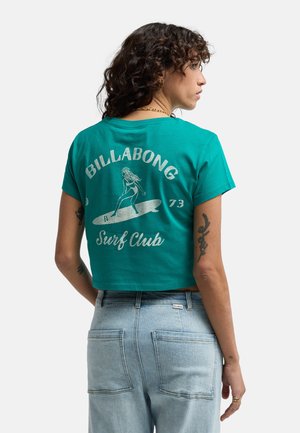 Kobieta o kręconych włosach, ubrana w skróconą turkusową koszulkę Billabong Surf Club i jasnoniebieskie jeansy, stojąca bokiem z widocznymi tatuażami na ramionach.