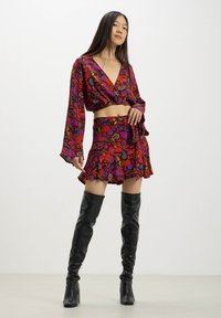 Blusa cropped a fantasia colorata e gonna coordinata in rosso, viola e verde. Presenta maniche lunghe, orlo svasato ed è indossata con stivali neri sopra il ginocchio.