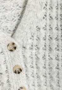 Cardigan lavorato a maglia grigio chiaro con un motivo tessurizzato, caratterizzato da grandi bottoni traslucidi lungo la parte anteriore. Dettaglio in tessuto morbido e peloso.