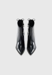 Une paire de bottines noires brillantes à bout pointu, avec des zips sur le côté et de petites anses de tirage, sur fond blanc.