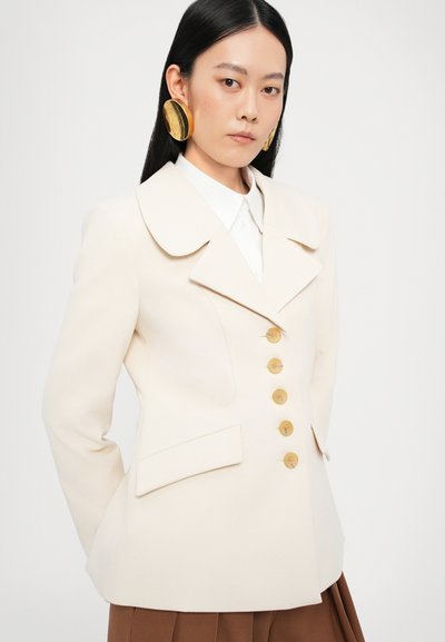 Blazer crema con un gran cuello, seis botones dorados, bolsillos laterales y un corte estructurado, usado sobre una camisa blanca y pantalones marrones.