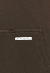 Tissu de sweat-shirt marron présentant une petite étiquette métallique argentée avec le texte "bazix republic" près de la couture de la poche. Texture lisse.