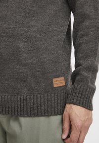 Pull en tricot gris foncé avec des poignets et un ourlet côtelés. Présente une étiquette en cuir avec "11 PROJECT" en bas à gauche. Texture douce.
