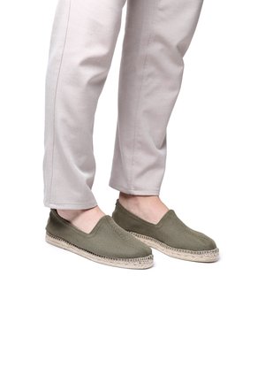 Green woven slip-on shoes with a jute rope sole, featuring a low-cut design and soft textile upper. Paired with light-colored pants.