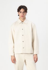 Lys beige, kravejakke lavet af tekstureret stof, med knaplukning foran og lange ærmer. Glat, minimalistisk design.