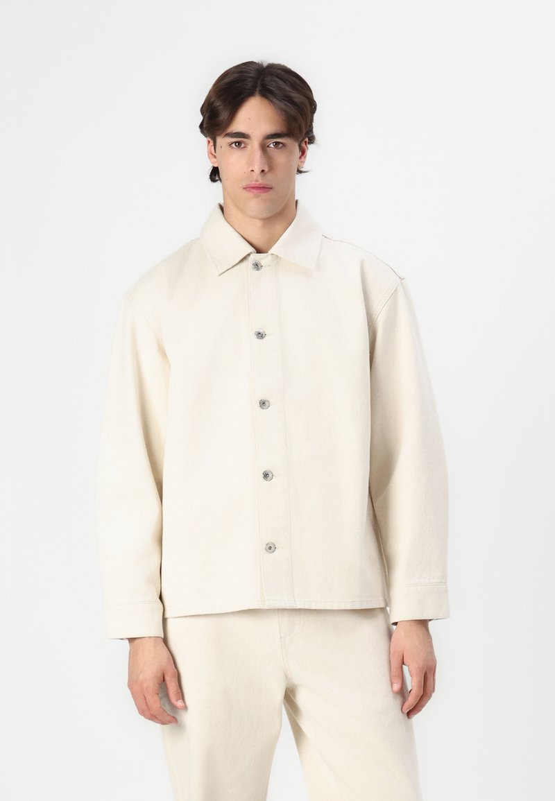 Lys beige, kravejakke lavet af tekstureret stof, med knaplukning foran og lange ærmer. Glat, minimalistisk design.