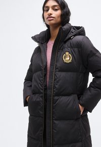 Μαύρο μπουφάν puffer με φερμουάρ μπροστά και κουκούλα, με χρυσό έμβλημα στο στήθος. Ομαλή υφή με σχέδιο καπιτονέ και πλαϊνές τσέπες.