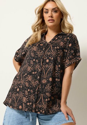 Vrouw die een zwart blouse met knopen draagt, versierd met perzikkleurige bloemenpatronen, en lichtblauwe denim shorts, poserend tegen een effen achtergrond.