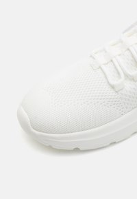Bílé sportovní boty s texturovanou síťovinou, polstrovanou podrážkou a plochými tkaničkami. Design má nenápadný, minimalistický styl.