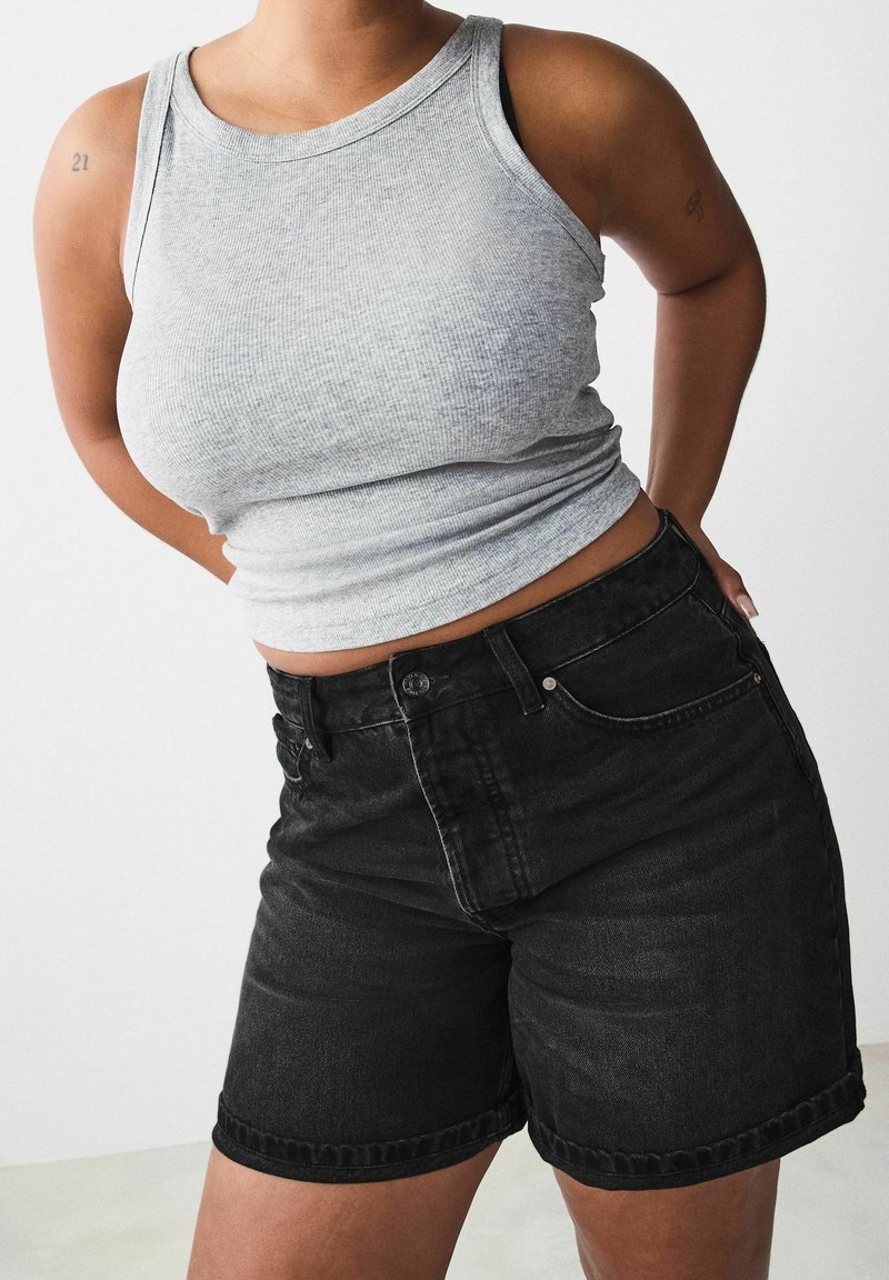 Personne portant un débardeur côtelé gris sans manches et un short en denim noir, debout les mains derrière le dos.