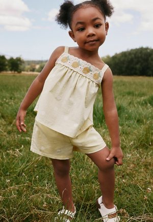 Niña joven con camiseta amarilla sin mangas y pantalones cortos, parada sobre el césped, sosteniendo su pierna, con árboles y cielo nublado en el fondo.
