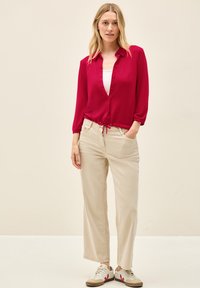 Himbeerfarbene Bluse mit Knöpfen und Kordelzugsaum, über einem weißen Shirt getragen; kombiniert mit beigen Hosen und Sneakers mit roten Akzenten.