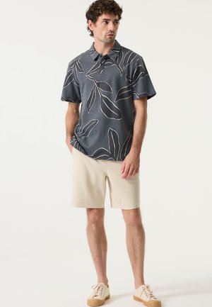 Korte mouwen polo in marineblauw met wit bladmotief, gemaakt van gestructureerde stof. Gecombineerd met lichtbeige shorts en casual sneakers.