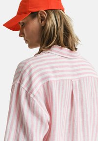 Camicia a righe rosa e bianca con vestibilità ampia, colletto e dettaglio con piega sul retro, realizzata in un tessuto leggero. Presenta un'etichetta visibile.