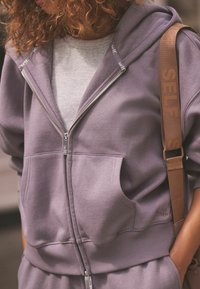 Fialová zip-up mikina s veľkými prednými vreckami, rebrovanými manžetami a spodným lemom, s hnedým pásom s nápisom "SELF" a svetlosivou vnútornou vrstvou.