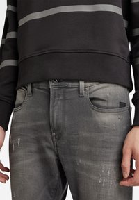 Primer plano de unos jeans grises desgastados con detalles de desgaste y una sudadera negra con rayas horizontales en gris claro en las mangas y el pecho.