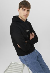 Zwarte hoodie met logo aan de voorkant, capuchon met trekkoorden en geribde manchetten, gecombineerd met lichtblauwe spijkerbroek. Model poseert tegen een witte achtergrond.