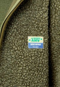 Veste en polaire zippée vert olive avec une doublure intérieure douce et texturée, et une étiquette indiquant "LA MAISON AMV SUPER VINTAGE."