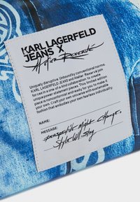 Tela de mezclilla azul con una etiqueta distintiva que presenta texto negro y escritura a mano. La etiqueta incluye nombres de marcas y un mensaje sobre la individualidad.