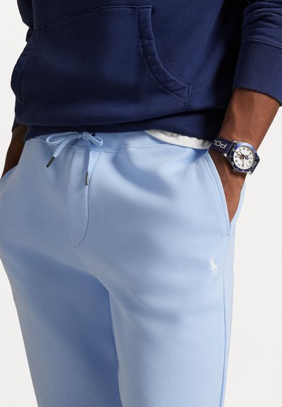 Lyst blå sweatpants med snor i taljen, med et lille hvidt logo på benet og et struktureret stof. Mørk navy hoodie delvist synlig.