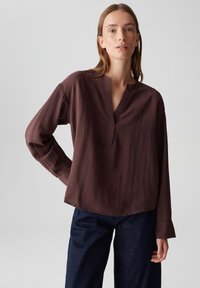 Woman wearing a loose, long-sleeve brown blouse with a split neckline and dark blue jeans, standing with one hand on hip.