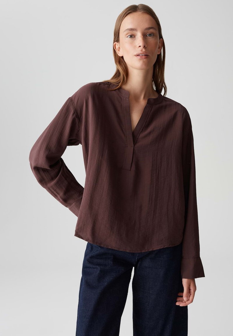 Woman wearing a loose, long-sleeve brown blouse with a split neckline and dark blue jeans, standing with one hand on hip.