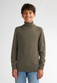 Maglione a collo alto lavorato a maglia in verde oliva, con maniche lunghe e polsini a coste, abbinato a jeans blu chiaro. Tessuto semplice e texturizzato.