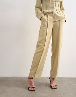 Osoba oblečená v béžových mierne priliehavých nohaviciach a zodpovedajúcom saku, s rukami vreckách, stojaca na betónovej podlahe v čiernych sandáloch na podpätku.