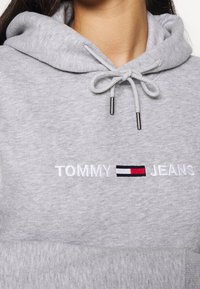Felpa grigia in pile con cappuccio regolabile, logo ricamato "TOMMY JEANS" e un accento rettangolare rosso, bianco e blu sotto il testo.
