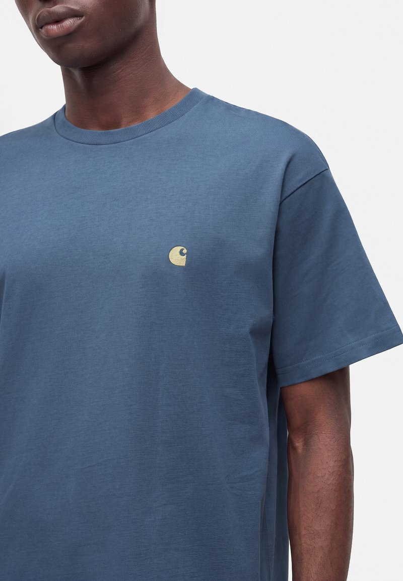 Mann trägt ein blaues kurzärmeliges T-Shirt mit kleinem gelbem Logo auf der linken Brust vor einfachem Hintergrund.
