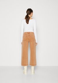 Pantalones de pana de pierna ancha en marrón claro con un acabado texturizado, que cuentan con bolsillos traseros y una cintura alta, combinados con una blusa blanca de manga larga.