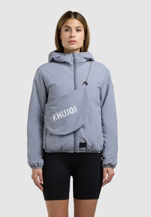 Frau trägt eine hellgraue Kapuzenjacke mit Reißverschluss, schwarze Radlerhose und eine Umhängetasche mit dem Schriftzug „KHUJO 2001“ auf der Vorderseite.