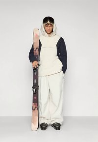 Ski outfit featuring a cream and navy jacket, white trousers, and black ski boots. Holding pink and brown skis with bindings. Black goggles worn.