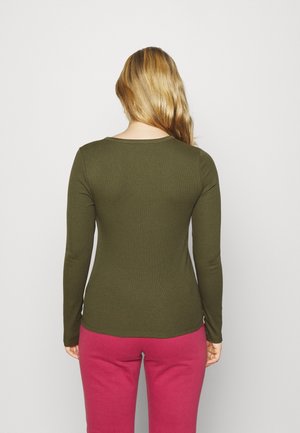 Femme aux longs cheveux blonds portant un haut à manches longues vert olive et un pantalon magenta, debout, dos tourné vers le spectateur sur un fond uni.
