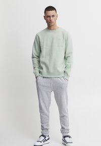 Felpa verde chiaro con polsini a coste, abbinata a pantaloni della tuta grigi con una cintura elastica. Le scarpe da ginnastica sono bianche con dettagli blu navy.