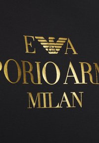 Černá látka s výraznou zlatou typografií. Text zahrnuje logo "E A" a "EMPORIO ARMANI MILAN," s texturovaným vzhledem písmen.